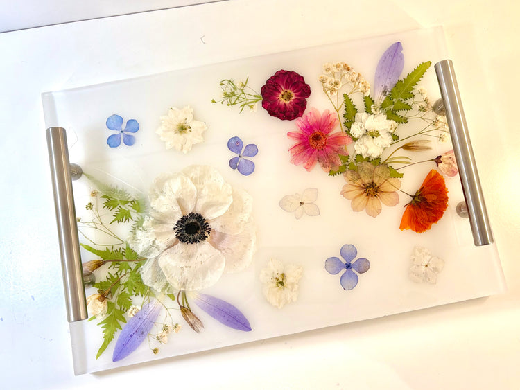 Decorative glass tray with pressed flowers on a white surface. Clear rectangular resin vanity tray with real embedded pink and white flowers and elegant silver handles.