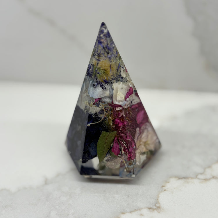 Pyramid-shaped crystal with embedded flowers and leaves on a marble surface. A large, multi-faceted diamond prism ring holder made from crystal clear resin with floating dried wildflowers inside.