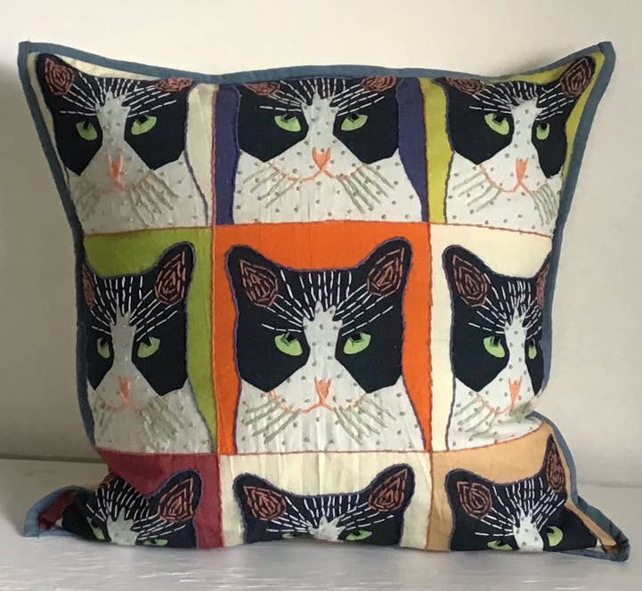 A square-shaped decorative pillow with an applique design featuring multiple cats in a multicolored patchwork pattern, with a mix of black, white, and orange colors.