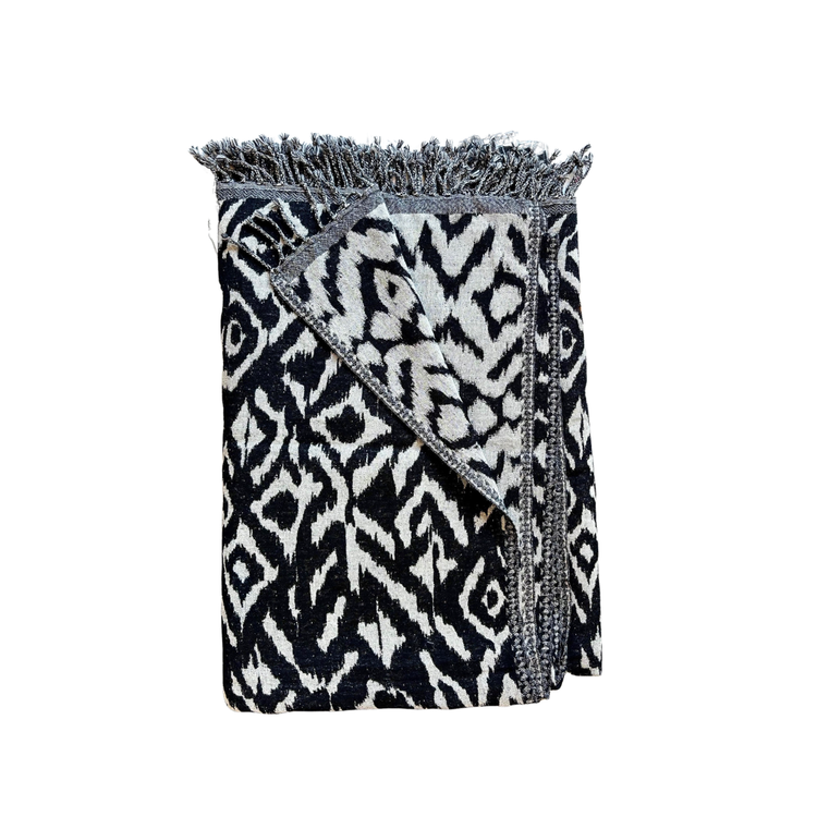 Folded blanket with black and white pattern on a white background. black and cream geometric Ikat patterned wool throw blanket with soft fringed edges, handloomed by village artisans.