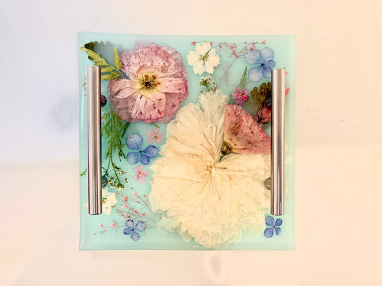 Floral design on a resin tray with two metal bookmarks on a light background. Square clear resin tray featuring a beautiful arrangement of embedded dried yellow and red flowers with metallic accent handles.