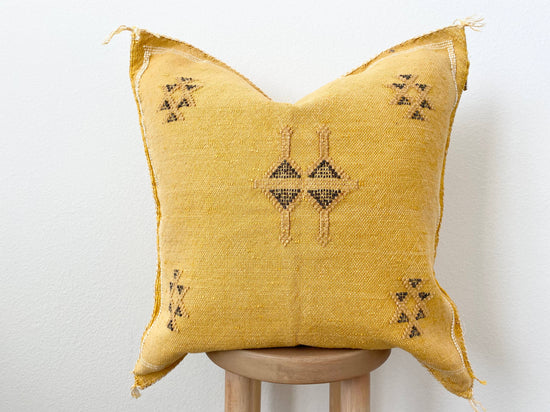 An orange/yellow silk sabra pillow with geometric patterns, displayed on a wooden stool against a white background.