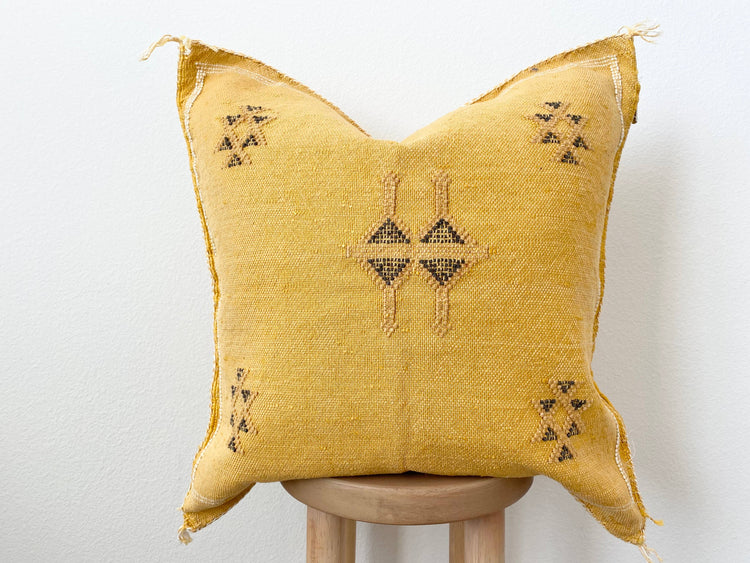 An orange/yellow silk sabra pillow with geometric patterns, displayed on a wooden stool against a white background.