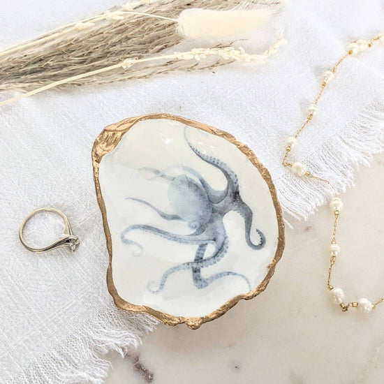 A handcrafted oyster shell ring dish with gold leaf trim, painted with a blue octopus design, displayed on a white surface accompanied by a gold ring and a pearl necklace.